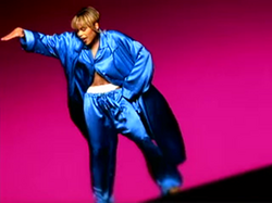 A women showing a white boxer inside her pants, with her right hand up and her left hand on her hip, backed by a dark pink background.