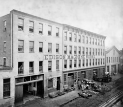 A perspective shot in black and white of a building with the Edison Machine Works name painted in the center. Several windows are missing. An old fashioned garage appears on the closest section on the left. A group of people stands in front of the single room central office with a pile of construction material.
