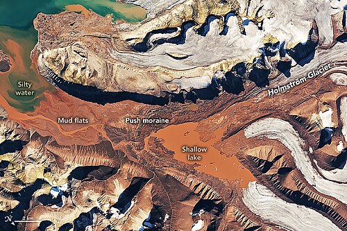 Late-summer satellite view of Spitsbergen, showing the Holmstrom Glacier and meltwater pools stained deep-red by glacial silt, from erosion of soft, iron-rich Devonian sediments