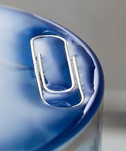 This paper clip is under the water level, which has risen gently and smoothly. Surface tension prevents the clip from submerging and the water from overflowing the glass edges.