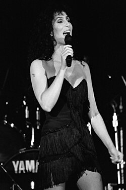 Cher sings on stage in a black fringed mini dress with spaghetti straps, her dark curly hair framing her face. A drum set is visible in the background.