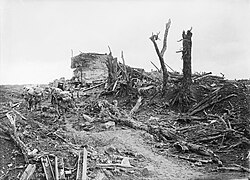 Heavily laden soldiers trudge through mud past the shattered remains of a concrete structure. Around them broken trees, steel beams and other pieces of debris have been strewn.