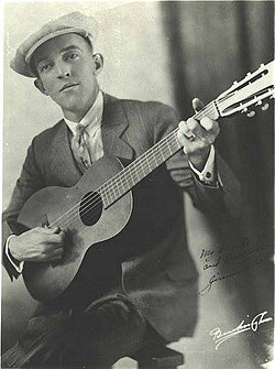 Autographed black and white picture of a man (Jimmie Rodgers) dressed in a striped suit, a white shirt, and wearing a tie and a beret. Sitting down, he holds the guitar to his chest resting the body on the right side and raising the neck while playing a C chord