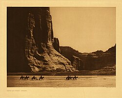 Navajo. Seven riders on horseback and dog trek against background of canyon cliffs. Edward S. Curtis (1904)