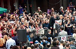 A large crowd of onlookers behind barriers surrounding a platform, with Scarlett Johansson in a black and white dress and emcee Leron Gubler in a black suit on the platform and smiling to the crowd