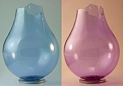 A neodymium glass light bulb, with the base and inner coating removed, under two different types of light: fluorescent on the left, and incandescent on the right.