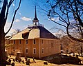 A large square church stands in behind a cemetery. The church has a small turret in the center where a bell is exposed to view.